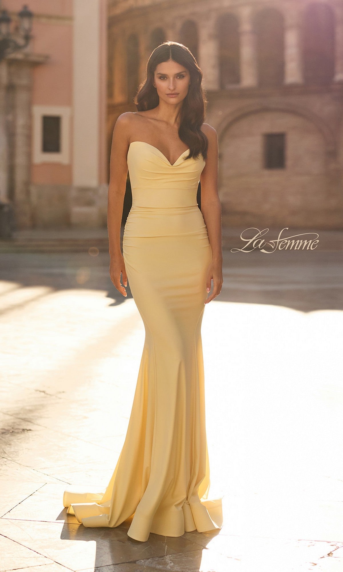 A woman stands outdoors in front of historic architecture, wearing the La Femme 33864 long prom dress in pale yellow—a fitted, strapless jersey gown with a slight train.